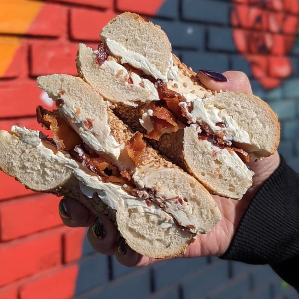 A hand holds a Stella Blue Bistro sesame bagel with cream cheese and bacon against a brick wall.
