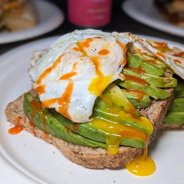 Avocado toast with a fried egg, runny yolk, and hot sauce on a white plate.